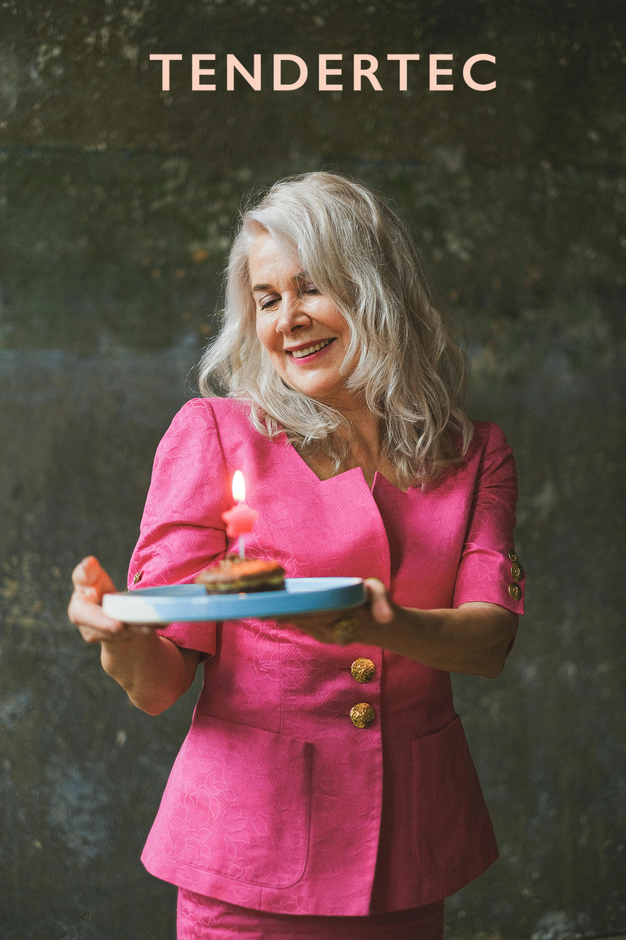 Older women wearing a pink blazer and holding a plate with a small round cake that has a lit candle